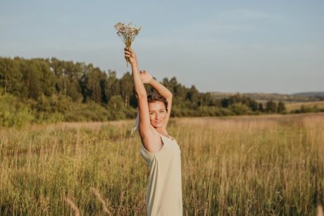 A joyful woman in a white dress holds wildflowers in a sunny field, embracing the serene summer outdoors.